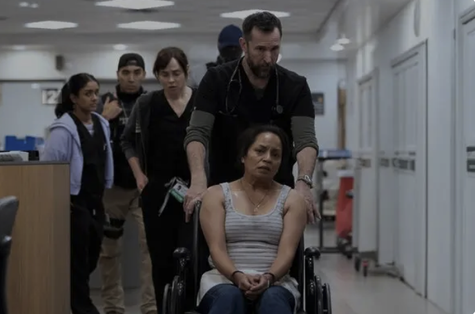 A  doctor wheels a woman in a wheelchair through a hospital E.R. Behind him are staffers and I.C.E. agents.